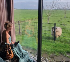 Eelke zit voor het raam met een zachte groene deken omgeslagen en een gezichtsmasker op. Ze kijkt lachend naar een schaap buiten in een weiland, welke ook naar haar kijkt.