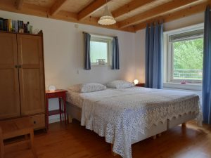 Tweepersoonsbed met sprei, nachtkastjes en twee ramen met blauwe gordijnen. Houten kledingkast