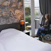 Eelke staat in haar rolstoel bij de openslaande deuren en doet het gordijn open, naast het bed. Ze kijkt naar buiten naar een klein terras met tafeltje en gele stoeltjes