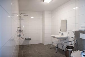 witte tegels aan de wand, grijze tegels op de grond, regendouche met zitje, onderijdbare wastafel en toilet met beugels