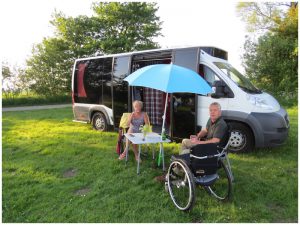 man in rolstoel met vrouw op campingstoel zitten voor een camper met groot glazen wand op een grasveld onder een blauwe parasol