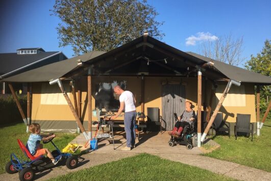 Eelke ligt in haar rolstoel voor de tent in de zon, haar neefje komt aanrijden op de skelter en zwager maakt de BBQ aan