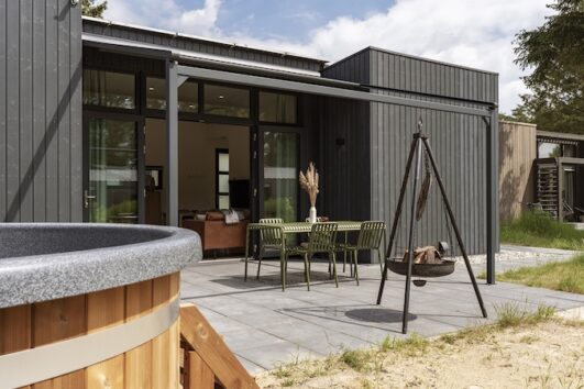 Zwart houten huisje, voorkant met terras en driepoot met vuurschaal (andere hoek)