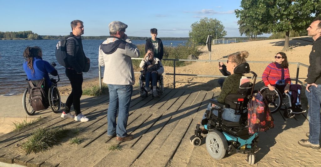 Groep journalisten staat op een houten vlonder bij een groot blauw meer en zandstrand