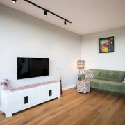 Hoek in de woonkamer met een fluweel groene bank, houten vloer, en witte lage kast met tv erboven