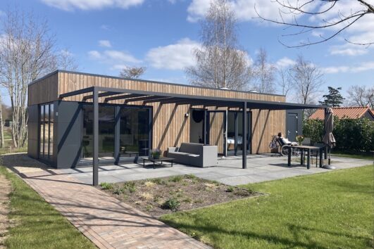 Houten langwerpig huisje met zwart/grijze kozijnen grote schuifdeuren. Een pergola boven het terras met kleine bank en eettafel met stoelen. Op de voorgrond een grasveld