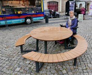 Eelke zit aan een ronde rolstoeltoegankelijke picknicktafel op een plein met foodtrucks achter haar