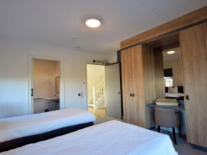 kamer met twee van elkaar staande bedden. De deur naar de aangepaste (ensuite) badkamer staat open en in de kamer is een grote kast met bureau en daarboven een spiegel