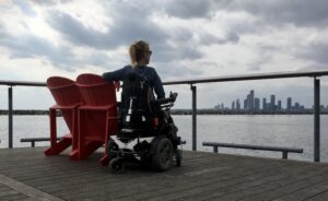 Eelke zit in haar rolstoel bij twee rode stoelen en kijkt naar het water met daarachter de skyline van Toronto