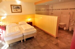 twee boxspringbedden, tegels op de vloer, licht gele muren. Open verbinding naar de badkamer me halfhoge muur als scheiding. toilet zichtbaar.