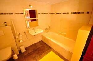 Badkamer met bad (vrij lage instap), onderrijdbare wastafel, met twee spiegels boen elkaar, stukje toilet met opklapbare beugel zichtbaar