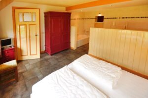 twee boxspringbedden, een rode kledingkast, tegels op de vloer, licht gele muren. Open verbinding naar de badkamer me halfhoge muur als scheiding. Bad zichtbaar.